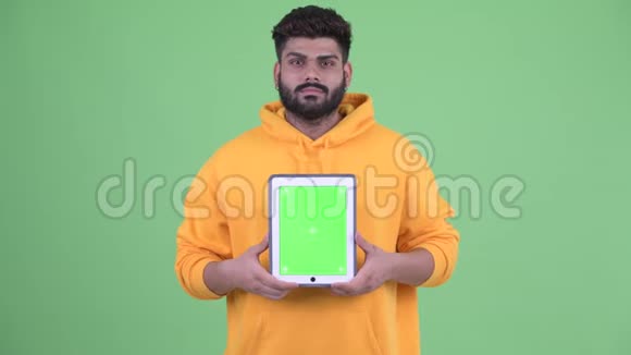 快乐年轻体重超重留胡子的印度男人一边说话一边展示数字平板电脑视频的预览图