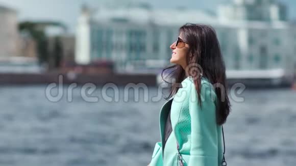 可爱的女孩吹着头发欣赏城市景观站在河堤上放松户外视频的预览图