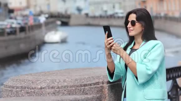 微笑时尚女游客用智能手机站在河上的桥上拍照视频的预览图