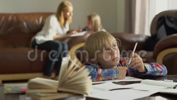 体贴的小男孩坐在桌子旁前景是笔记本而他的母亲和妹妹则坐在桌子旁视频的预览图