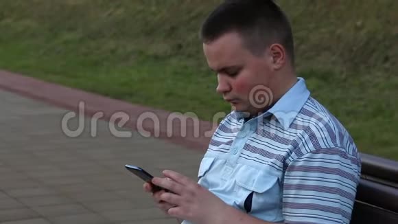 一个年轻人坐在长凳上在火车站的站台上手里拿着智能手机看着屏幕视频的预览图