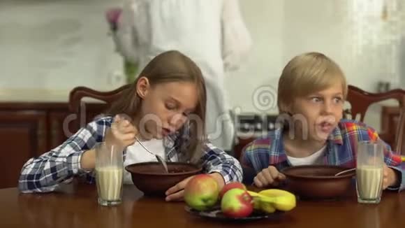 可爱的兄弟姐妹在厨房的餐桌上吃谷物和喝牛奶而母亲在厨房洗碗视频的预览图