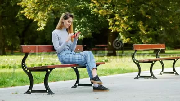 快乐的红发女孩在公园的长凳上用手机年轻女子在浏览互联网时微笑视频的预览图