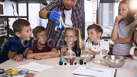 男韩语老师在课堂上展示青年学生化学实验玻璃瓶中蓝色化学反应视频的预览图
