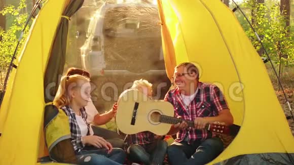 快乐的旅游家庭在阳光明媚的森林里的露营帐篷里玩得很开心友好的家庭笑着用吉他演唱歌曲视频的预览图