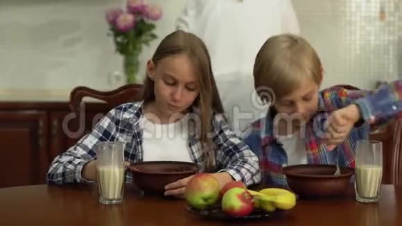 快乐的兄弟姐妹在厨房的餐桌上吃麦片和喝牛奶而母亲在厨房洗碗视频的预览图