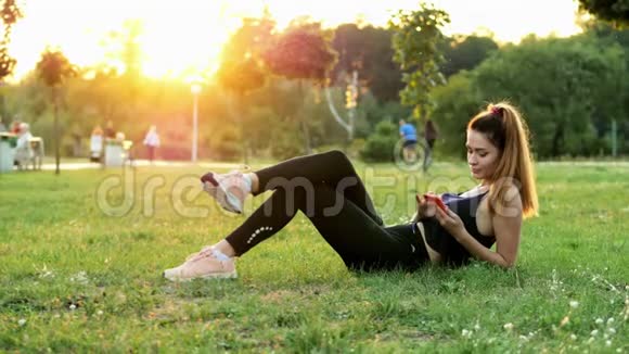 运动可爱的女人在日落时分躺在城市公园的草地上使用手机休息时间视频的预览图