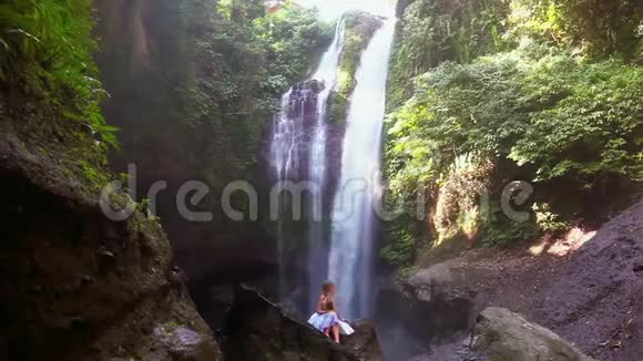 年轻女子坐在巴厘岛瀑布前的岩石上视频的预览图