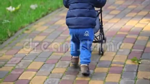 可爱的可爱的小学步男孩穿着蓝色便服在城市公园玩有趣的跑步和推玩具婴儿车视频的预览图
