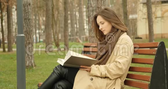 秋天公园里坐在长凳上的女人看书穿着棕色的壕沟和围巾阅读概念视频的预览图