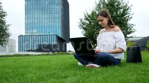 年轻的黑发女人坐在草地上用手提电脑用键盘打字用现代商业玻璃做背景视频的预览图