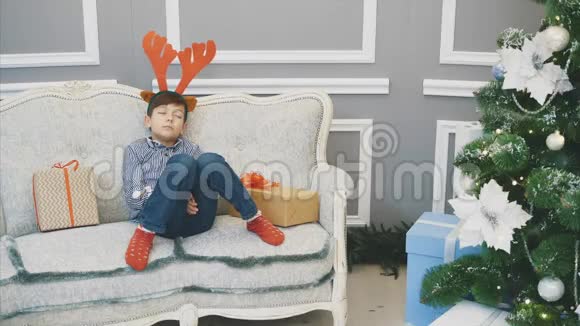 全身可爱的小男孩带着驯鹿头饰睡在靠近杉树的圣诞老人的沙发上视频的预览图