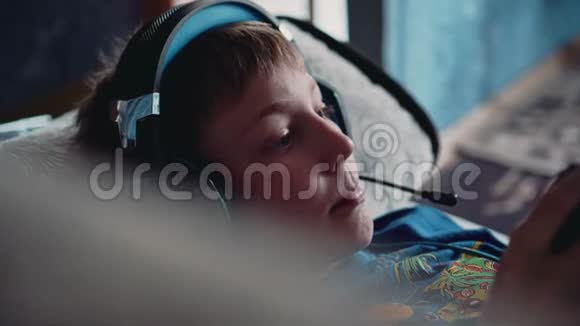 一个男孩头上戴着耳机躺在沙发上玩电子游戏他拿着控制器在家里很开心好心情休息视频的预览图