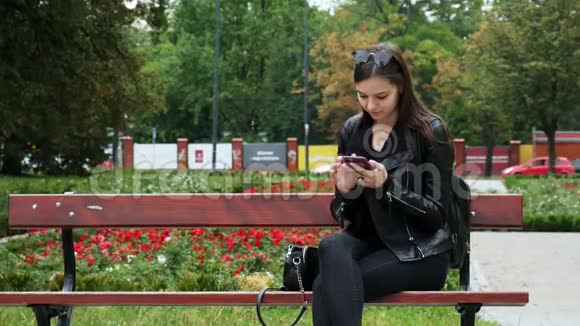 在公园里的长凳上一个年轻的女人在玩手机一个年轻的女人在上网时微笑视频的预览图