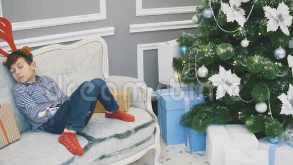 可爱的小男孩带着驯鹿头饰睡在靠近圣诞老人和冷杉树的沙发上视频的预览图