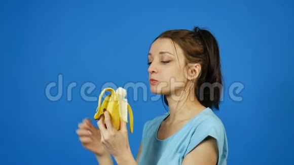 美丽的布鲁内特年轻女子手牵着黄色香蕉然后吃它在明亮的蓝色背景热带视频的预览图