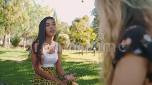 一位迷人的年轻女性在公园里坐在绿草上享受着与女性朋友交谈的乐趣视频的预览图