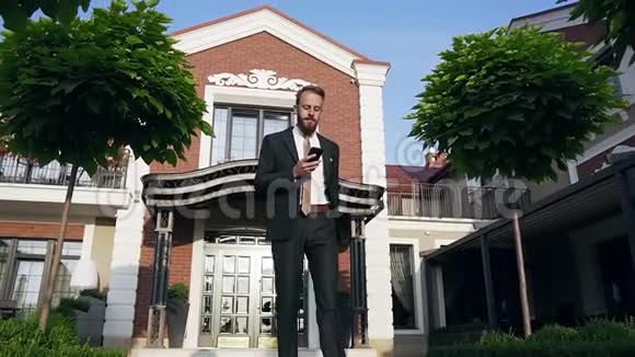 优雅修长的胡须男打电话走在美丽的大厦前的绿树之间视频的预览图