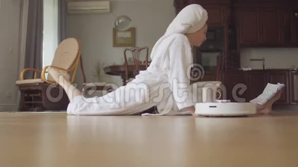 可爱的年轻灵活的白种人女孩淋浴后穿着浴衣坐在麻绳上阅读杂志喝咖啡或茶视频的预览图