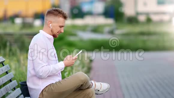 有魅力的时髦男性在耳机里放松坐在长凳上用智能手机听音乐视频的预览图