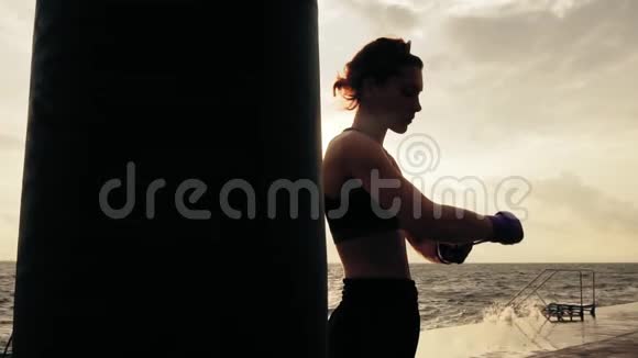 镜头轮动女拳击手在海边站在太阳旁边用拳击包裹手视频的预览图