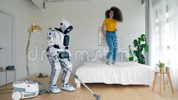 快乐的女孩跳到床上而机器人则打扫地板机器人半机器人和人类概念视频的预览图
