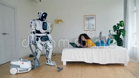 一个女人用笔记本电脑工作而一个机器人做真空清洁Cyborg和人类概念视频的预览图