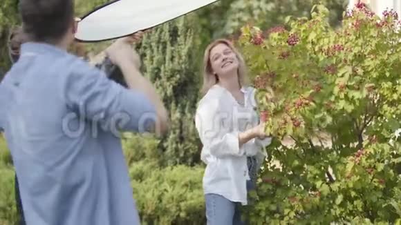 年轻积极的女孩穿着白色衬衫和牛仔裤在浆果灌木附近摆姿势为带相机的年轻人户外男性视频的预览图