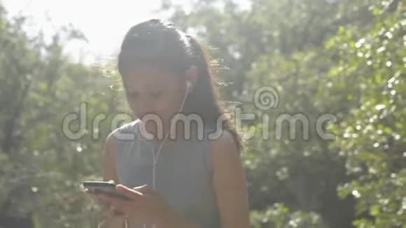 亚洲可爱的女孩使用手机带耳机早上站在红树林阳光下视频的预览图