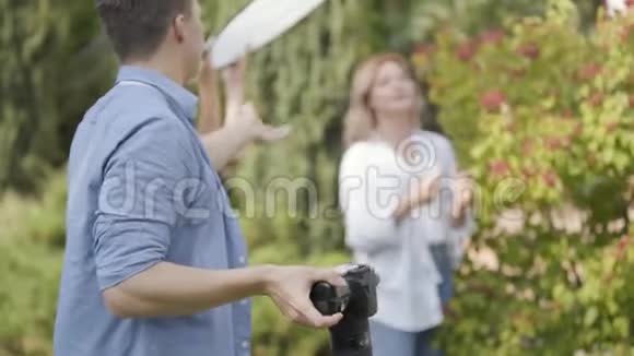 年轻积极的女孩穿着白色衬衫和牛仔裤在浆果灌木附近摆姿势为带相机的年轻人户外男性视频的预览图