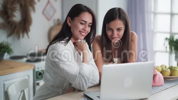 两个年轻的女人看着笔记本电脑讨论在厨房吃早餐视频的预览图
