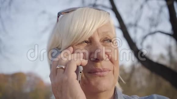 成熟的欧洲女人站在秋天公园用智能手机说话的特写肖像积极的高级人员视频的预览图