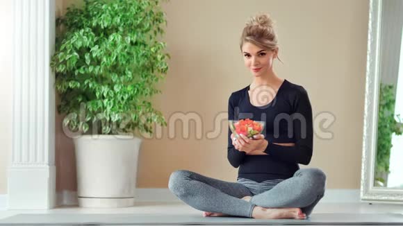 微笑的运动女人摆姿势坐在莲花的位置拿着盘子和新鲜蔬菜沙拉视频的预览图