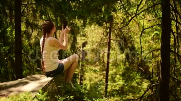 美丽的女孩坐在石头上自拍在智能手机上拍摄视频针叶林晴天视频的预览图