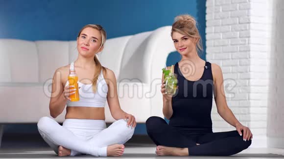 两位健身妇女摆出一副新鲜维生素鸡尾酒的姿势坐在瑜伽教室里摆着莲花姿势视频的预览图