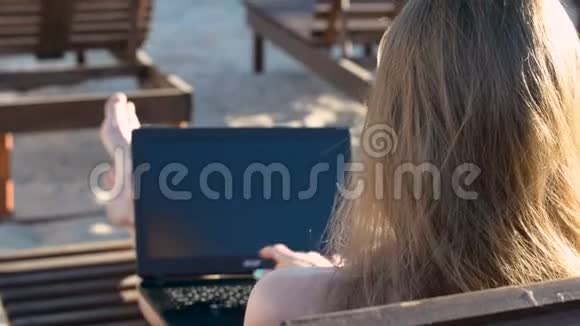 年轻女子坐在躺椅上在笔记本电脑上工作从后面看夏日视频的预览图