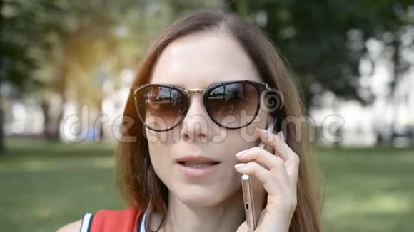 一个年轻漂亮的女孩戴着墨镜在夏天的公园里和朋友用手机聊天视频的预览图