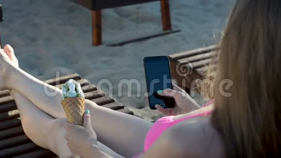 穿泳衣的女孩在海滩上坐在甲板椅上时使用智能手机手里拿着冰淇淋背面视频的预览图