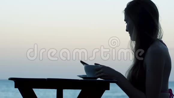 海滨咖啡馆里一位年轻女子的剪影手里拿着一杯茶和一部电话在智能手机上聊天视频的预览图