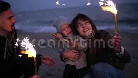 小女孩在父亲的怀里看着她的母亲父亲在黄昏的海滩上挥舞着火花年轻快乐视频的预览图