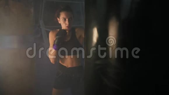 白种人强壮的女踢脚手在黑暗的健身房里用拳击袋和烟一起运动还和她一起踢拳击袋视频的预览图