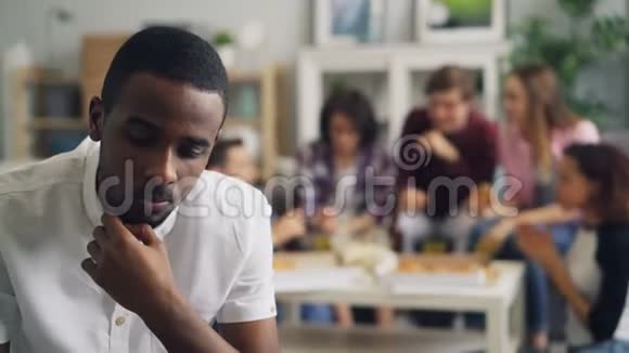 悲伤的非裔美国人一个人坐着开心的朋友在聚会上玩视频的预览图