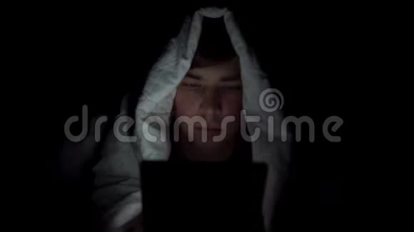 一个年轻人躺在毯子下面手里拿着一块药片黑暗中的人用垫子现代电子平板电脑视频的预览图
