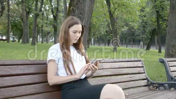 坐在长凳上的城市公园里一个快乐的女孩在用智能手机视频的预览图