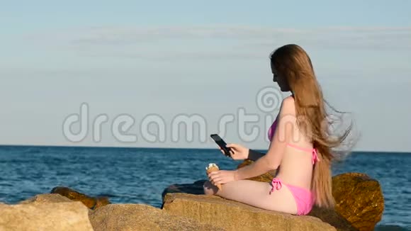 长发的年轻女子穿着泳衣坐在海边的岩石上打电话手里拿着冰淇淋你的哈风视频的预览图
