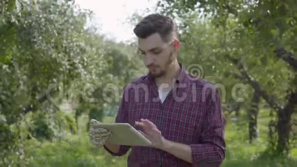 年轻自信的蓄着胡须的农民走过花园在平板电脑上打字环顾四周检查着视频的预览图
