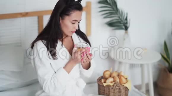 穿着浴袍的年轻女子在床上喝着茶吃着刚出炉的面包视频的预览图