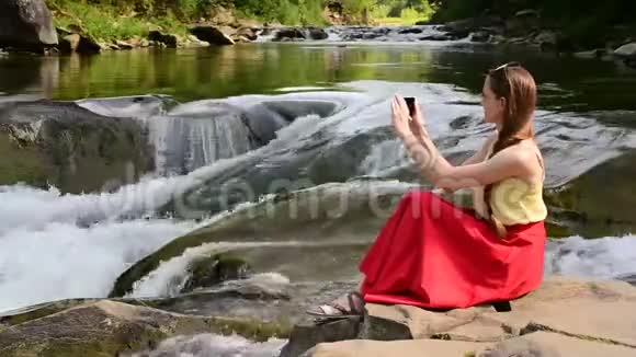 美丽的长发女孩穿着红色裙子坐在岩石上在山河瀑布的背景上自拍Summe视频的预览图