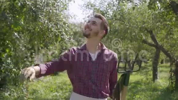 自信的年轻微笑着留着胡须的白种人农民拿着铁锹和鹳毛检查水果穿过花园视频的预览图