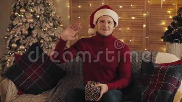 戴着圣诞帽和冬季毛衣的年轻帅哥坐在沙发上抱着礼物和视频的预览图
